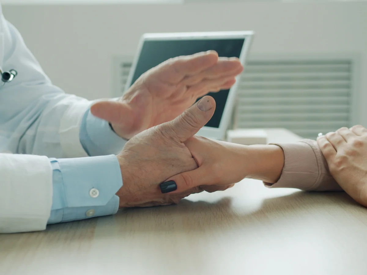 A clinician examines a person's hand and wrist across a desk.