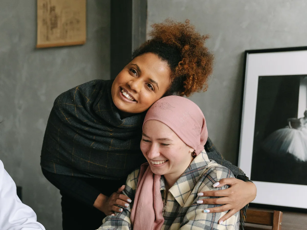 A woman in a head wrap smiles as a friend hugs her.