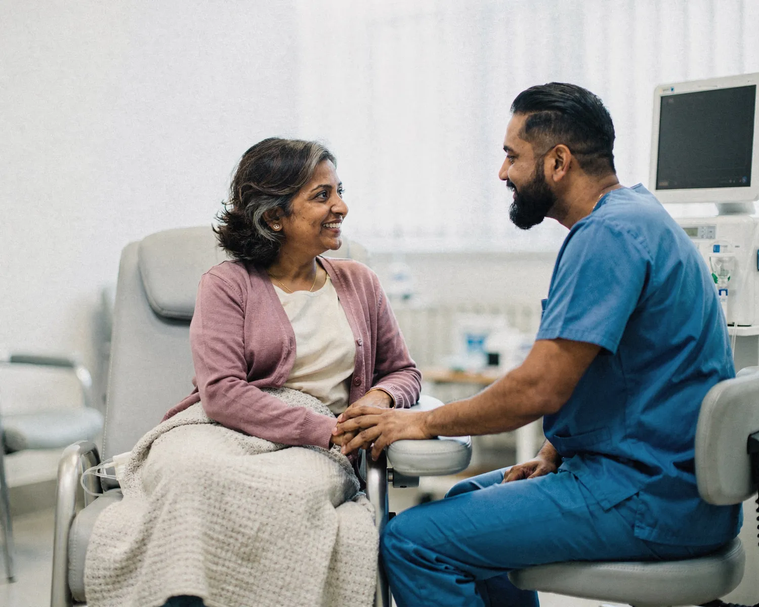A nurse holds a woman's hand and talks with her as she rests in a treatment chair at a dialysis center.