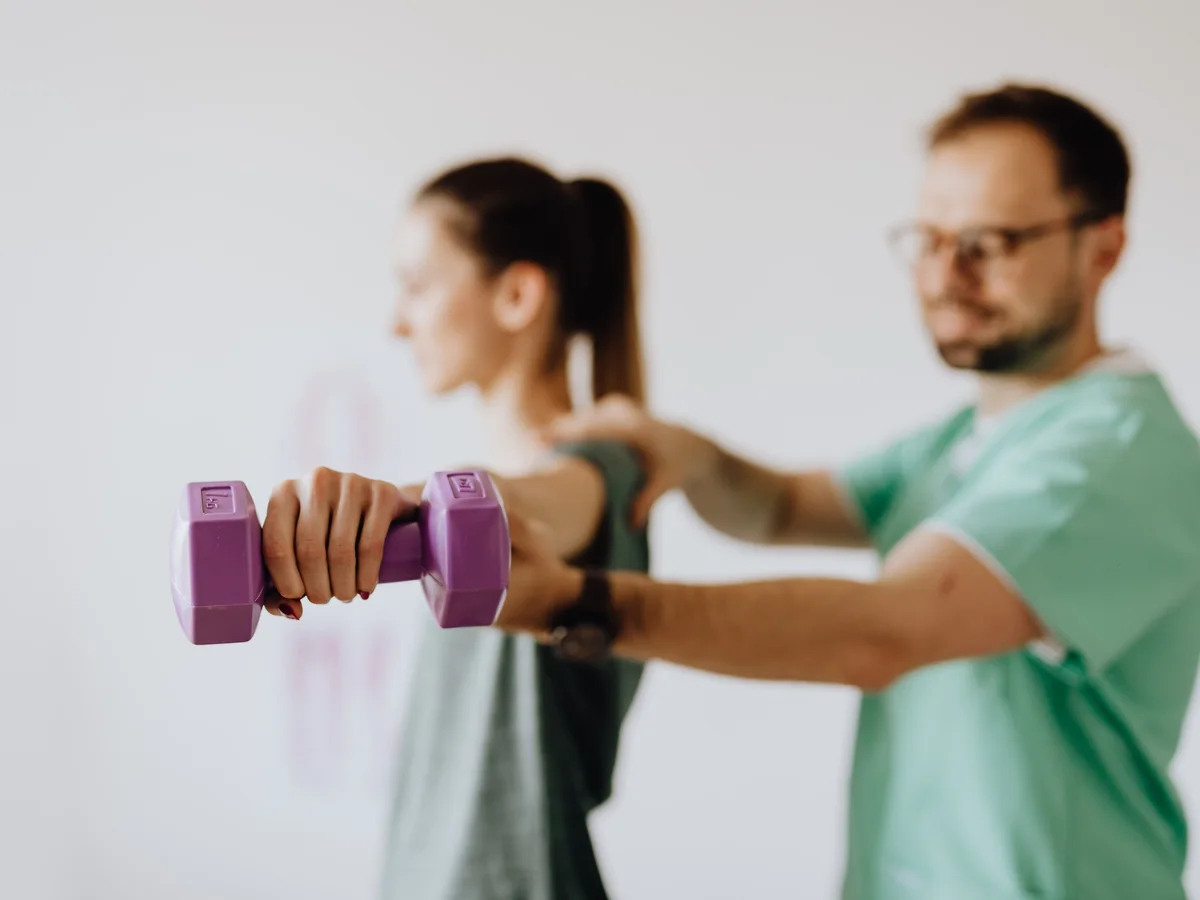 A therapist supports a woman during a shoulder exercise with a small weight.