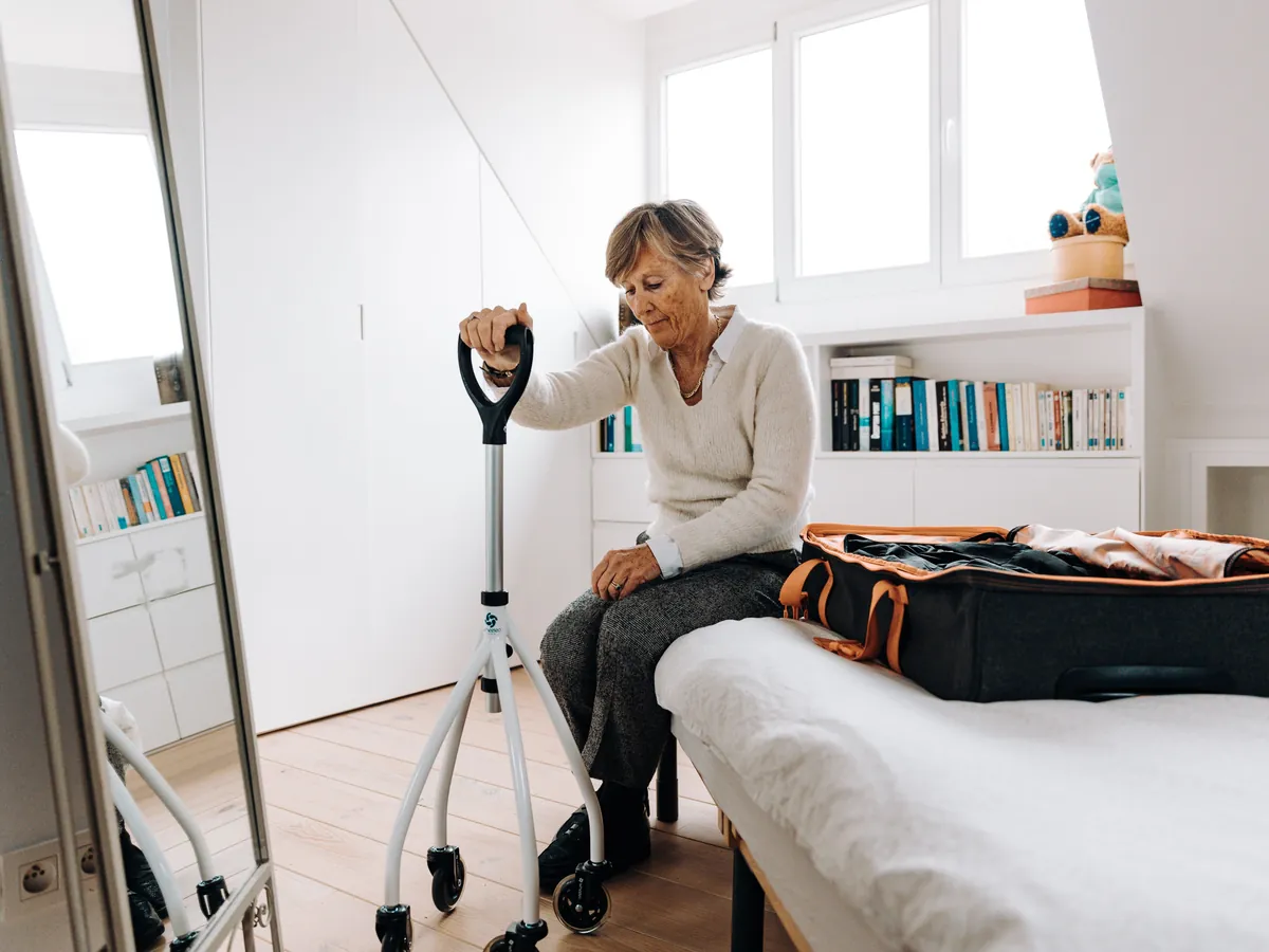 An older woman sits on her bed with a walking aid beside an open suitcase.