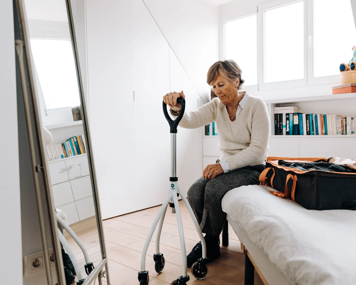 An older woman sits on her bed with a walking aid beside an open suitcase.
