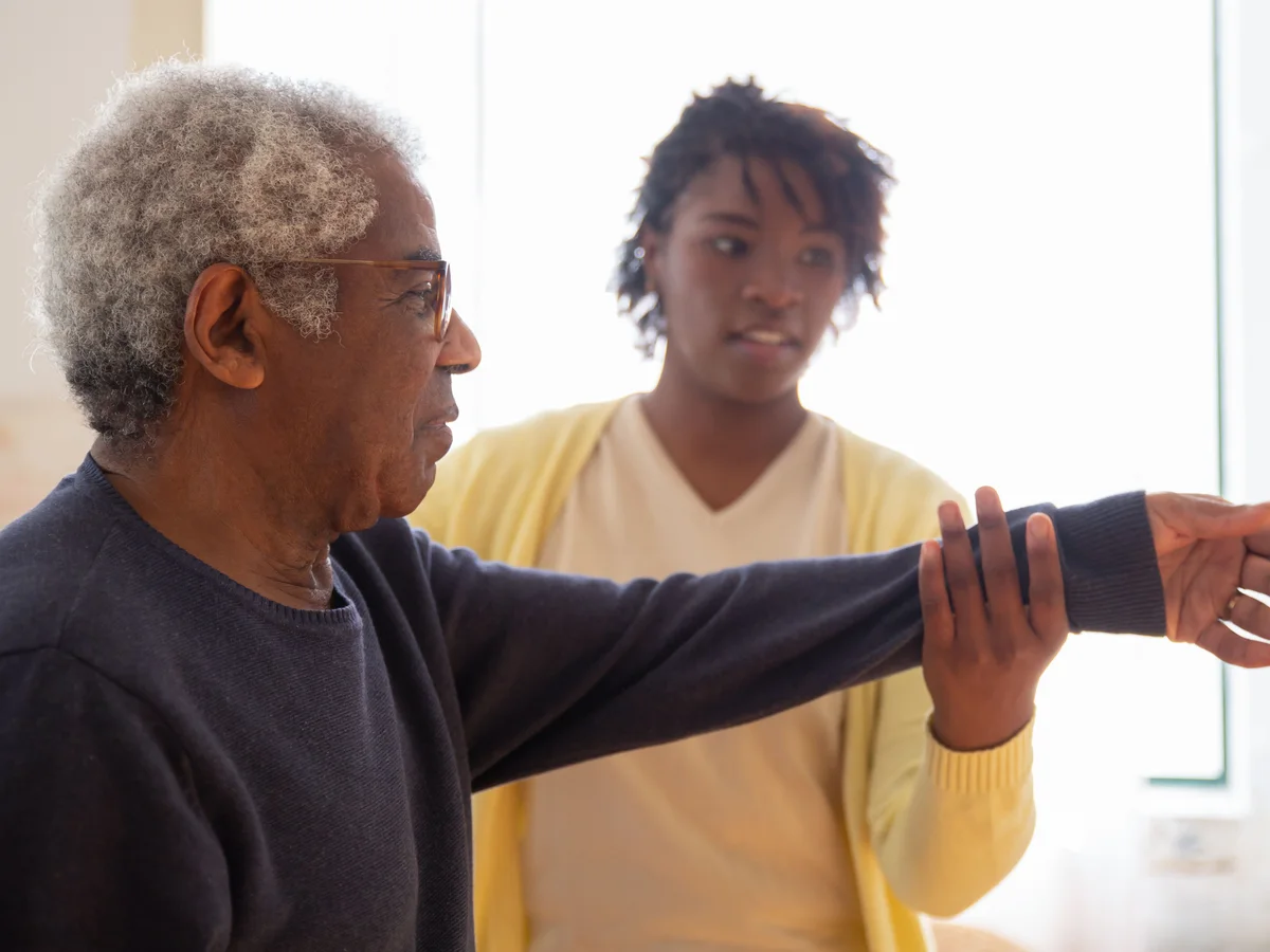 A woman supports an older man during an arm movement exercise.