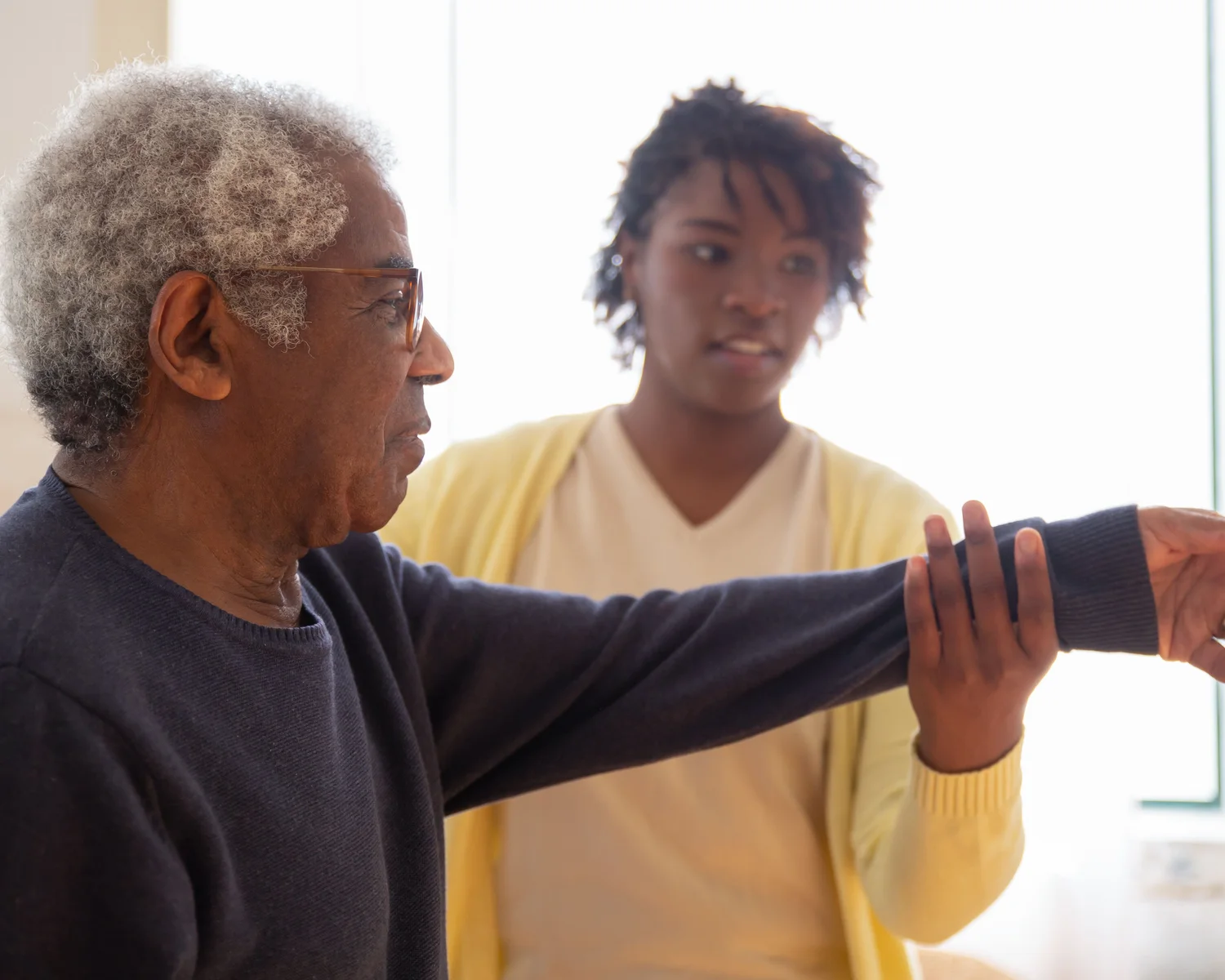A woman supports an older man during an arm movement exercise.
