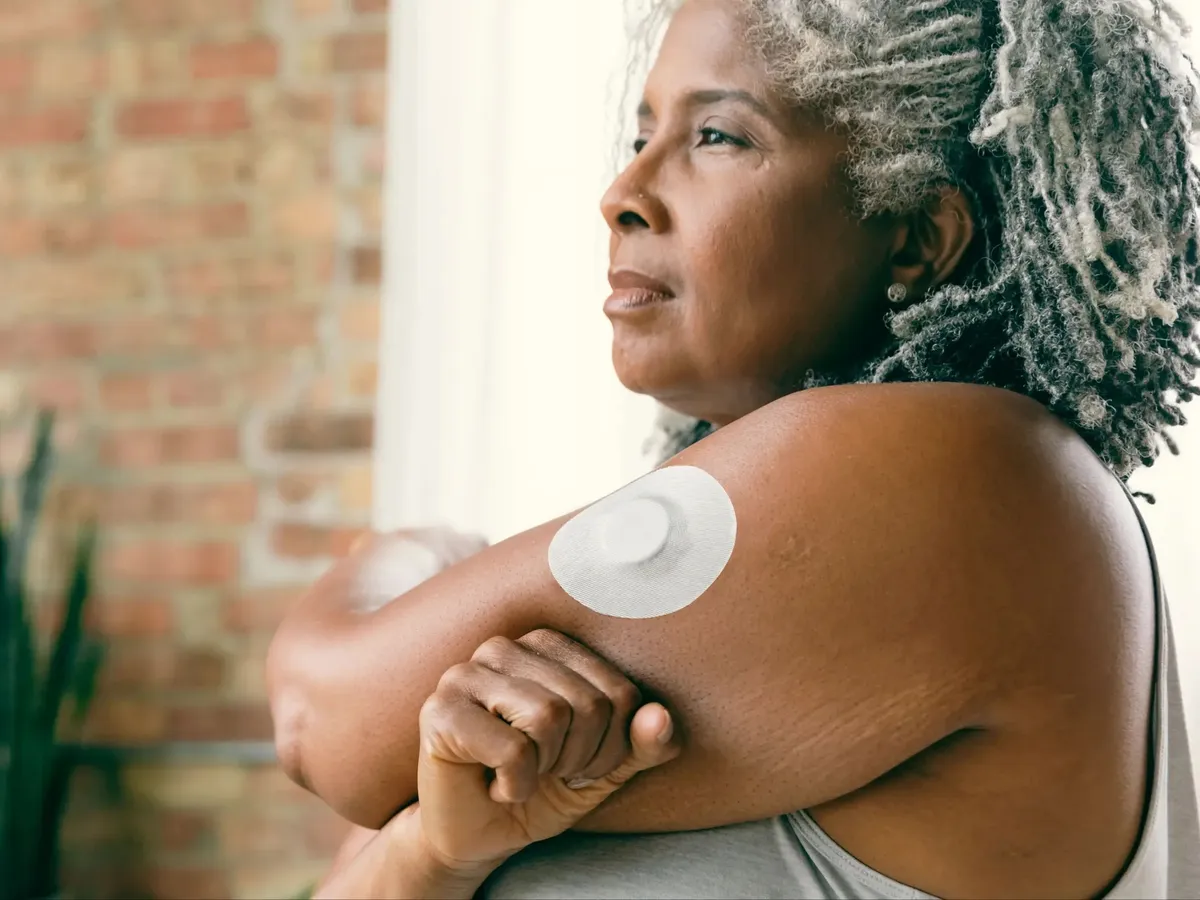 An older woman with a glucose sensor on her upper arm looks out of a window at home.