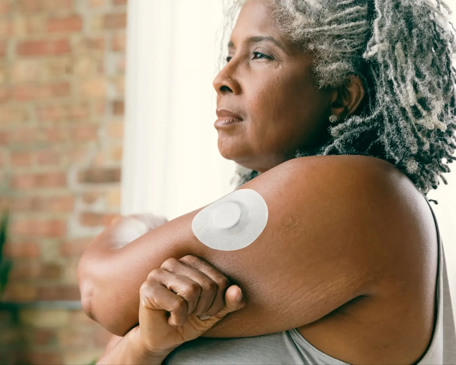An older woman with a glucose sensor on her upper arm looks out of a window at home.