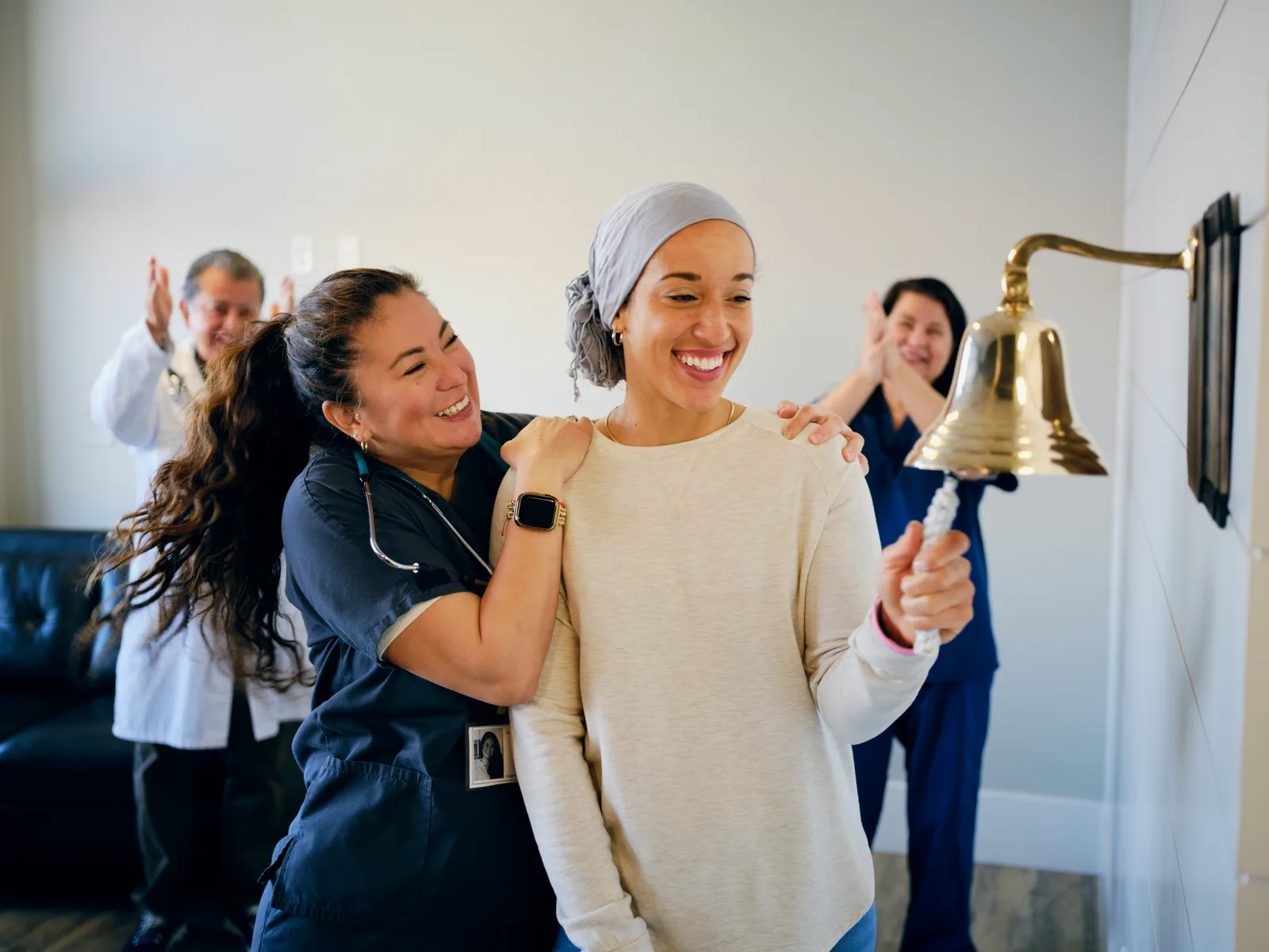 A smiling patient in a head wrap rings a brass bell while her nurse hugs her shoulders and two care team members applaud behind them.